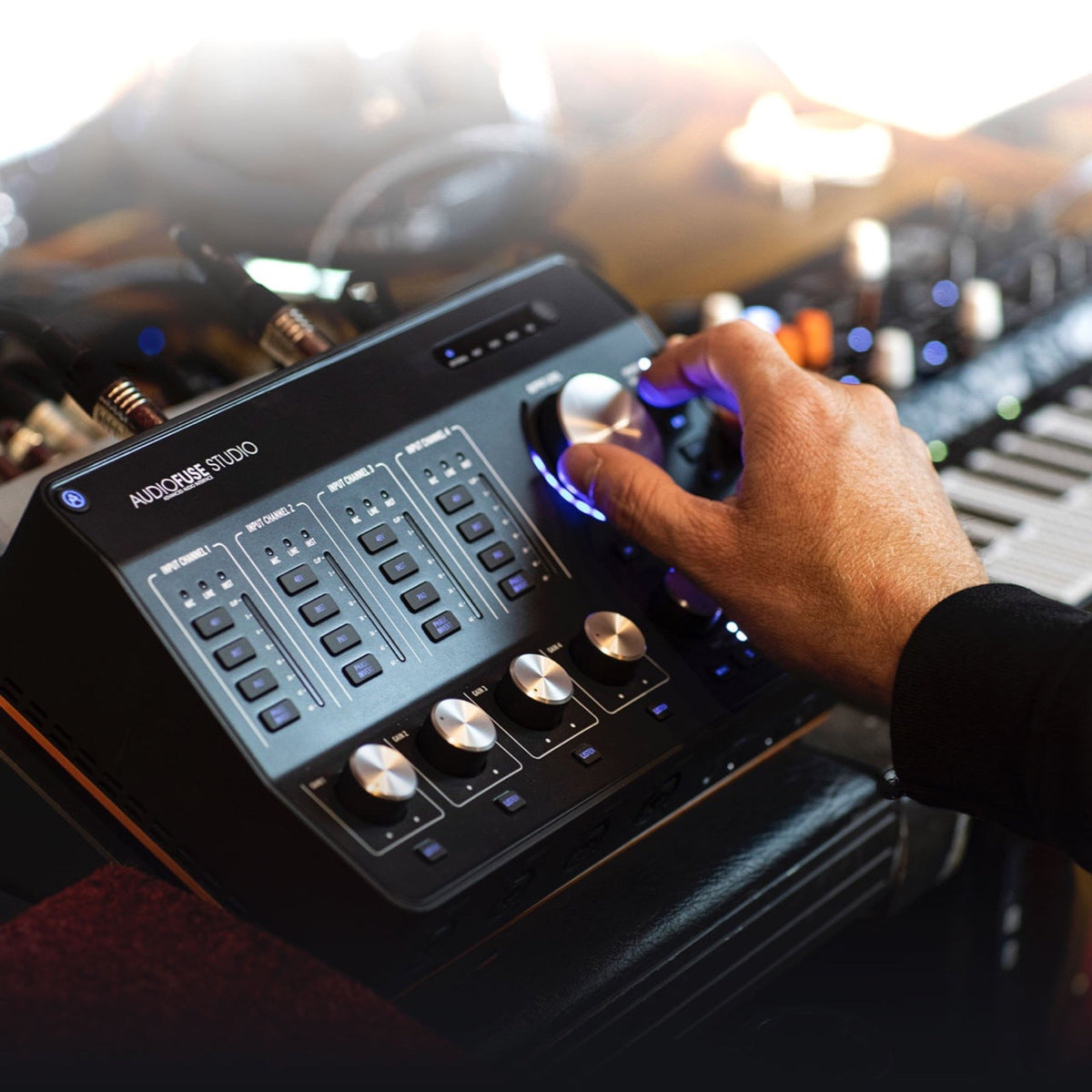 Arturia AudioFuse Studio on a studio desk with a hand on the monitor knob and a synthesizer behind it
